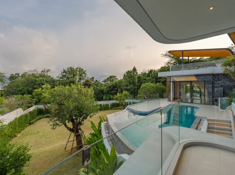 Modern house with a curved pool, glass railing, and a landscaped lawn area viewable from a balcony edge.