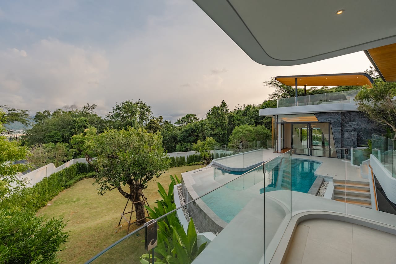 Modern house with a curved pool, glass railing, and a landscaped lawn area viewable from a balcony edge.