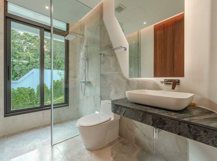 Modern bathroom with glass-enclosed shower, white vessel sink on a dark marble countertop, and a wall-mounted faucet; large window with greenery outside.