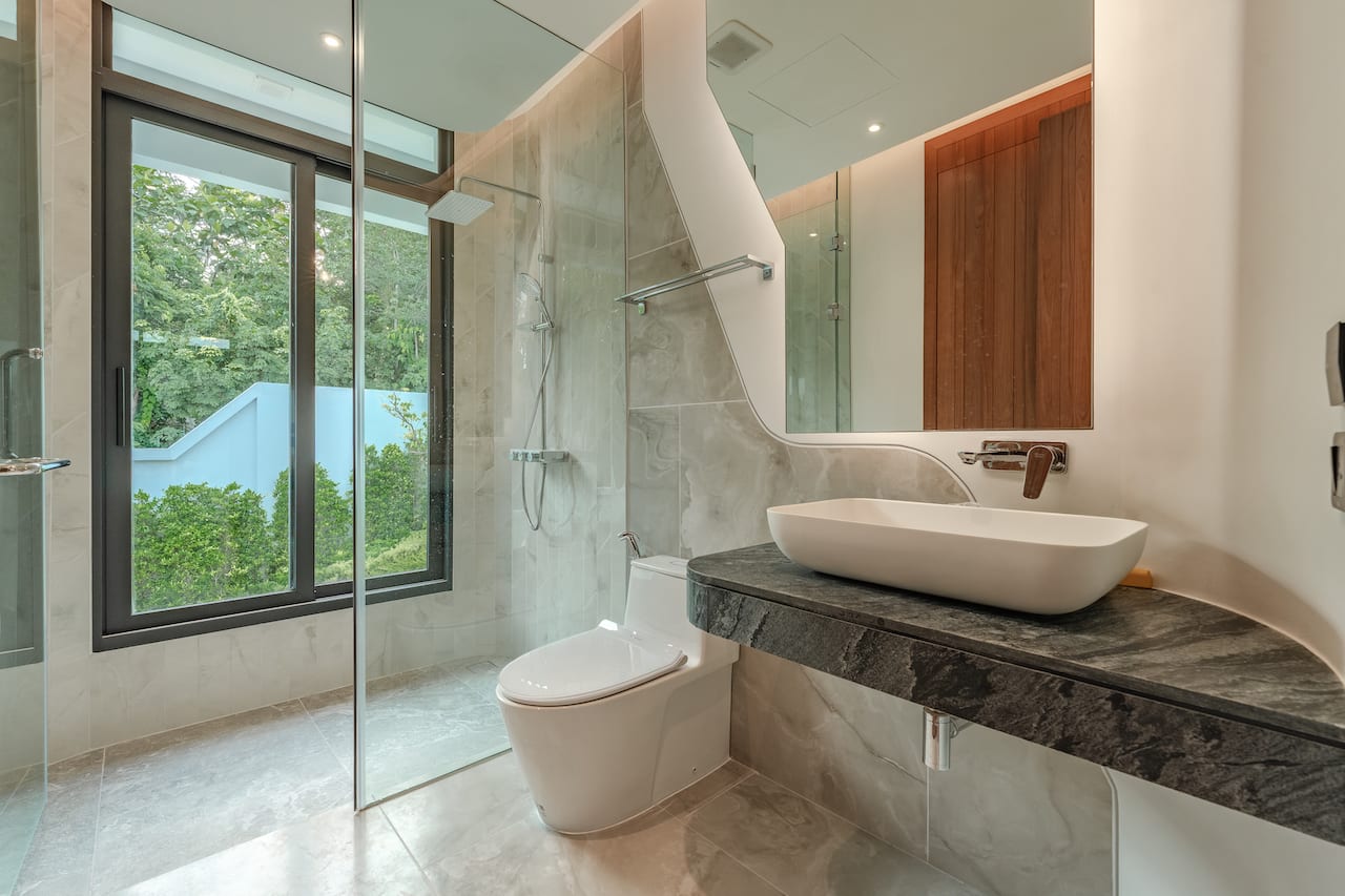 Modern bathroom with glass-enclosed shower, white vessel sink on a dark marble countertop, and a wall-mounted faucet; large window with greenery outside.