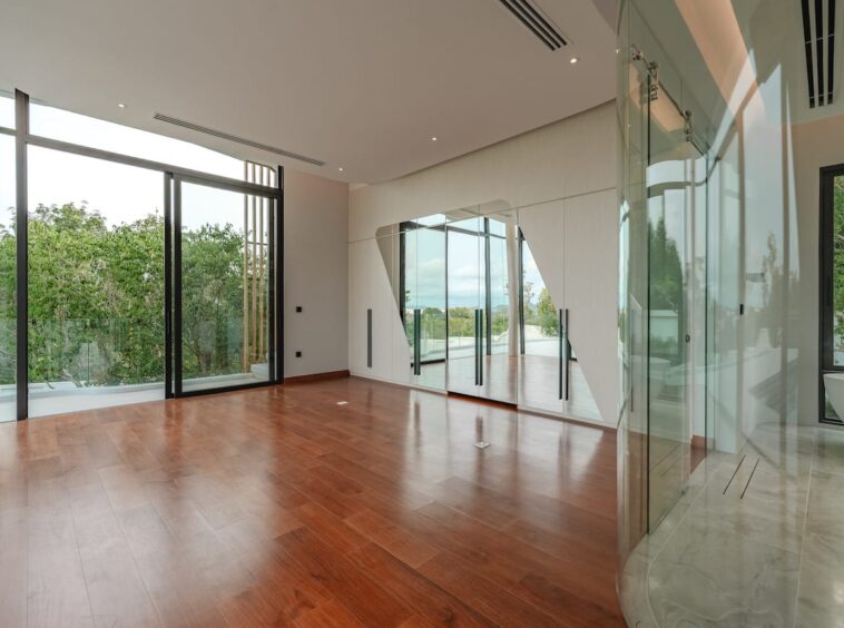 Spacious modern living area with hardwood floors, large glass doors to a balcony, and a mirrored wall reflecting greenery outside.