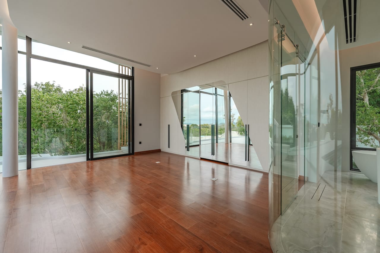 Spacious modern living area with hardwood floors, large glass doors to a balcony, and a mirrored wall reflecting greenery outside.
