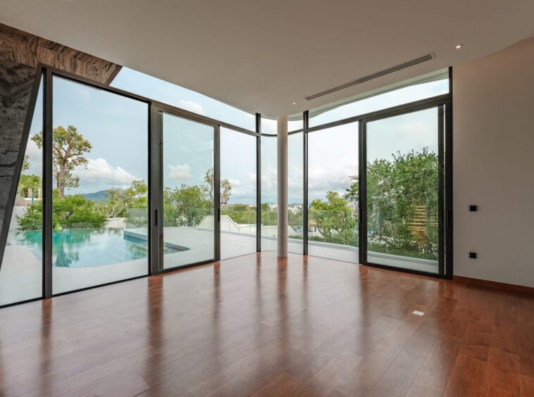 Spacious modern living area with floor-to-ceiling glass doors opening to a pool and garden view, wooden floors, and natural light.