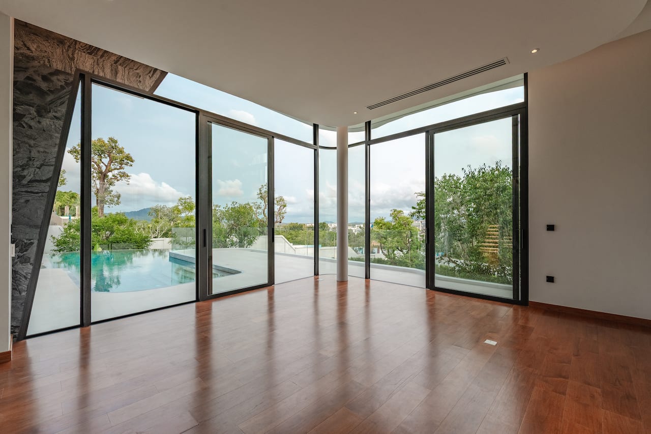 Spacious modern living area with floor-to-ceiling glass doors opening to a pool and garden view, wooden floors, and natural light.