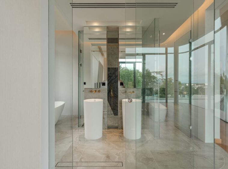 Bright modern bathroom with glass walls, two white pedestal sinks, a walk-in shower, a freestanding tub, and marble flooring.