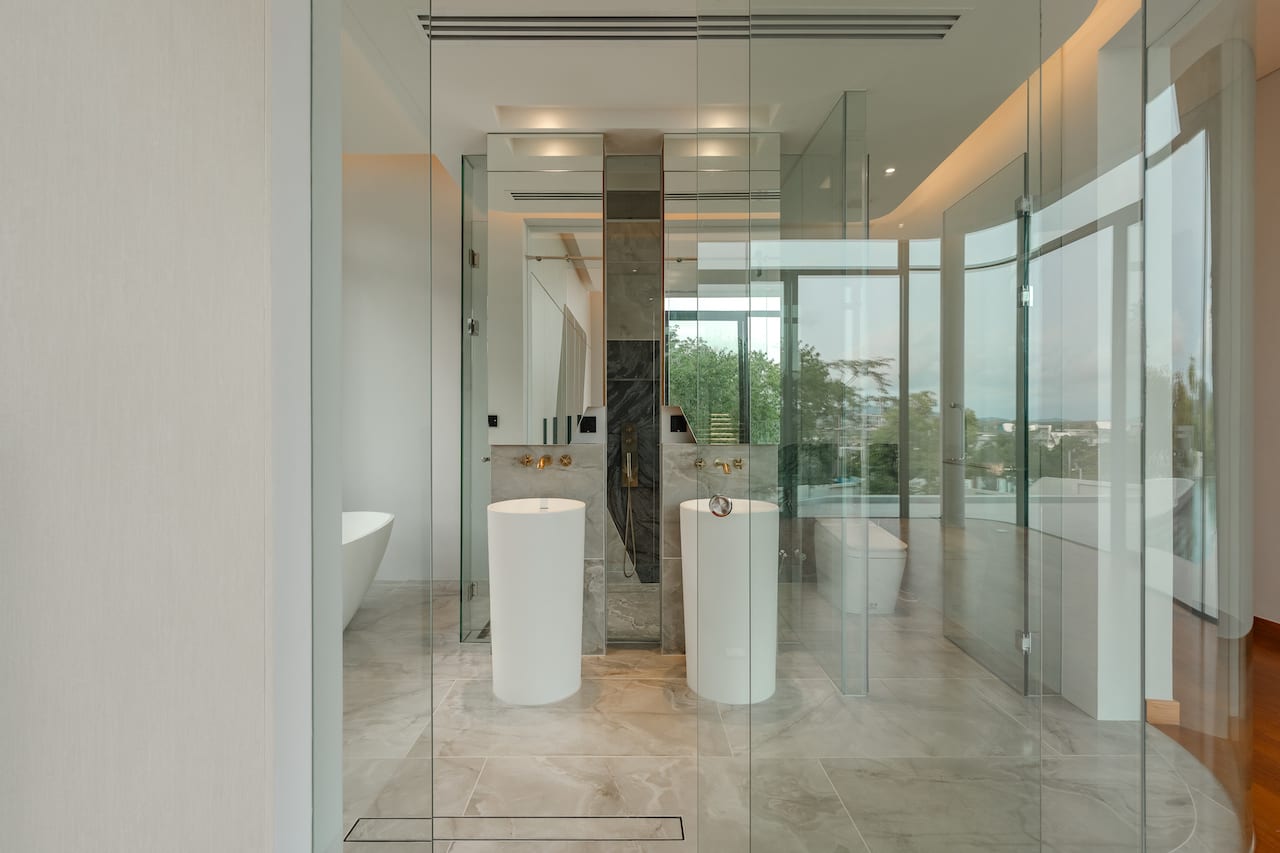 Bright modern bathroom with glass walls, two white pedestal sinks, a walk-in shower, a freestanding tub, and marble flooring.