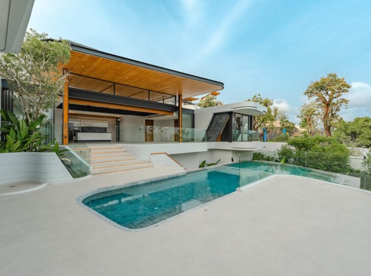 Luxurious modern villa with a glass-railed terrace, wooden ceiling accents, and a curved pool in the foreground.