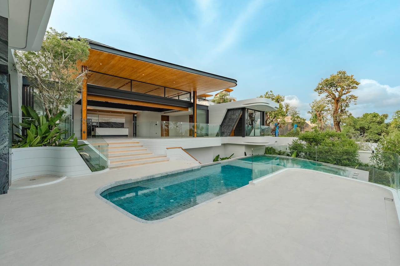 Luxurious modern villa with a glass-railed terrace, wooden ceiling accents, and a curved pool in the foreground.