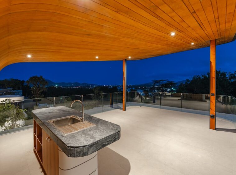 Night view terrace with a curved wooden roof, stone kitchen island with sink, and glass railing overlooking city lights at dusk.