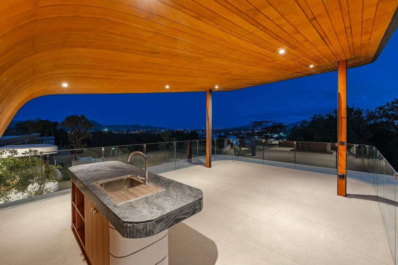 Night view terrace with a curved wooden roof, stone kitchen island with sink, and glass railing overlooking city lights at dusk.