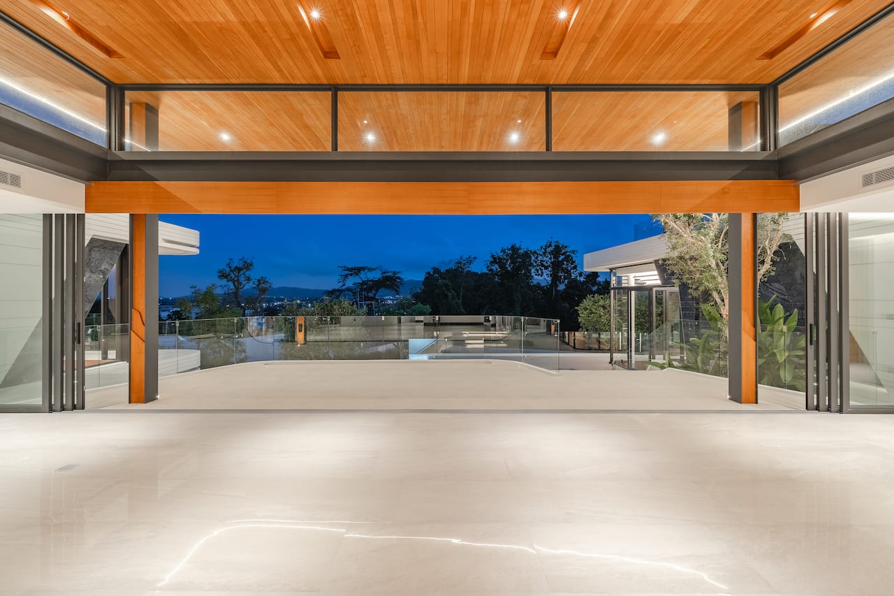 Open, minimalist living area with glass doors to a terrace, a polished white floor, and a warm wood ceiling. The night cityscape is visible beyond the glass railing.
