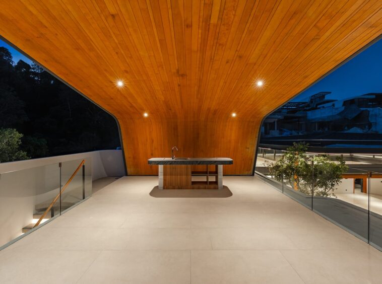 Architectural terrace with a wood-paneled curved ceiling, central island with sink, and glass railings at dusk.