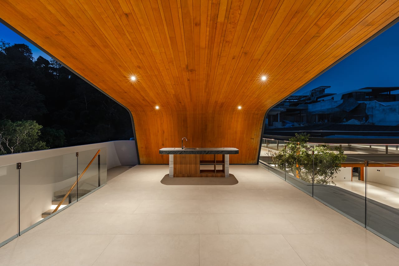 Architectural terrace with a wood-paneled curved ceiling, central island with sink, and glass railings at dusk.