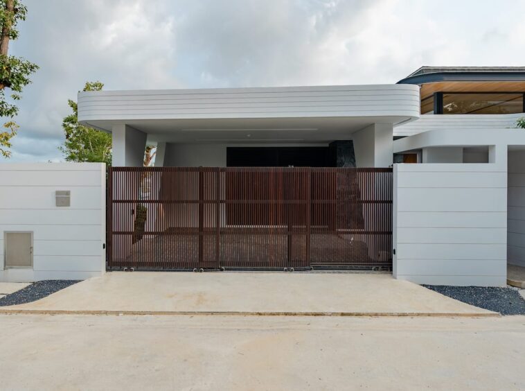 Modern white house with a tall brown vertical-slat gate and concrete driveway ahead of it, under a cloudy sky.