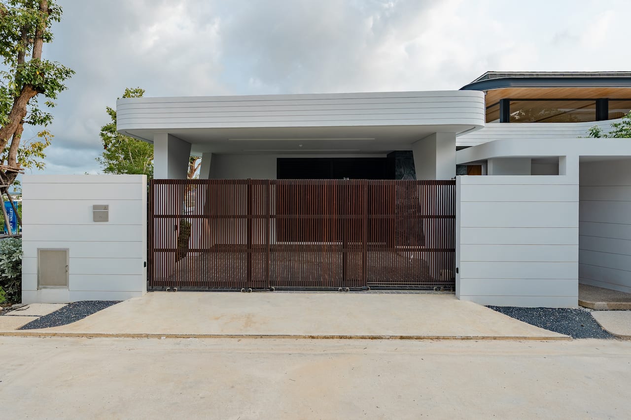 Modern white house with a tall brown vertical-slat gate and concrete driveway ahead of it, under a cloudy sky.