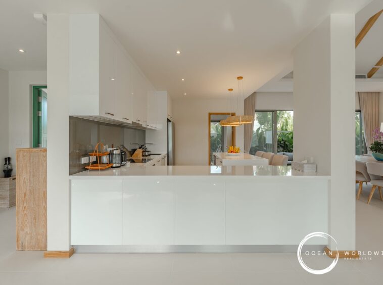 Bright, modern kitchen with white cabinetry and an island, open to a dining area with beige chairs and large windows showing greenery outside.