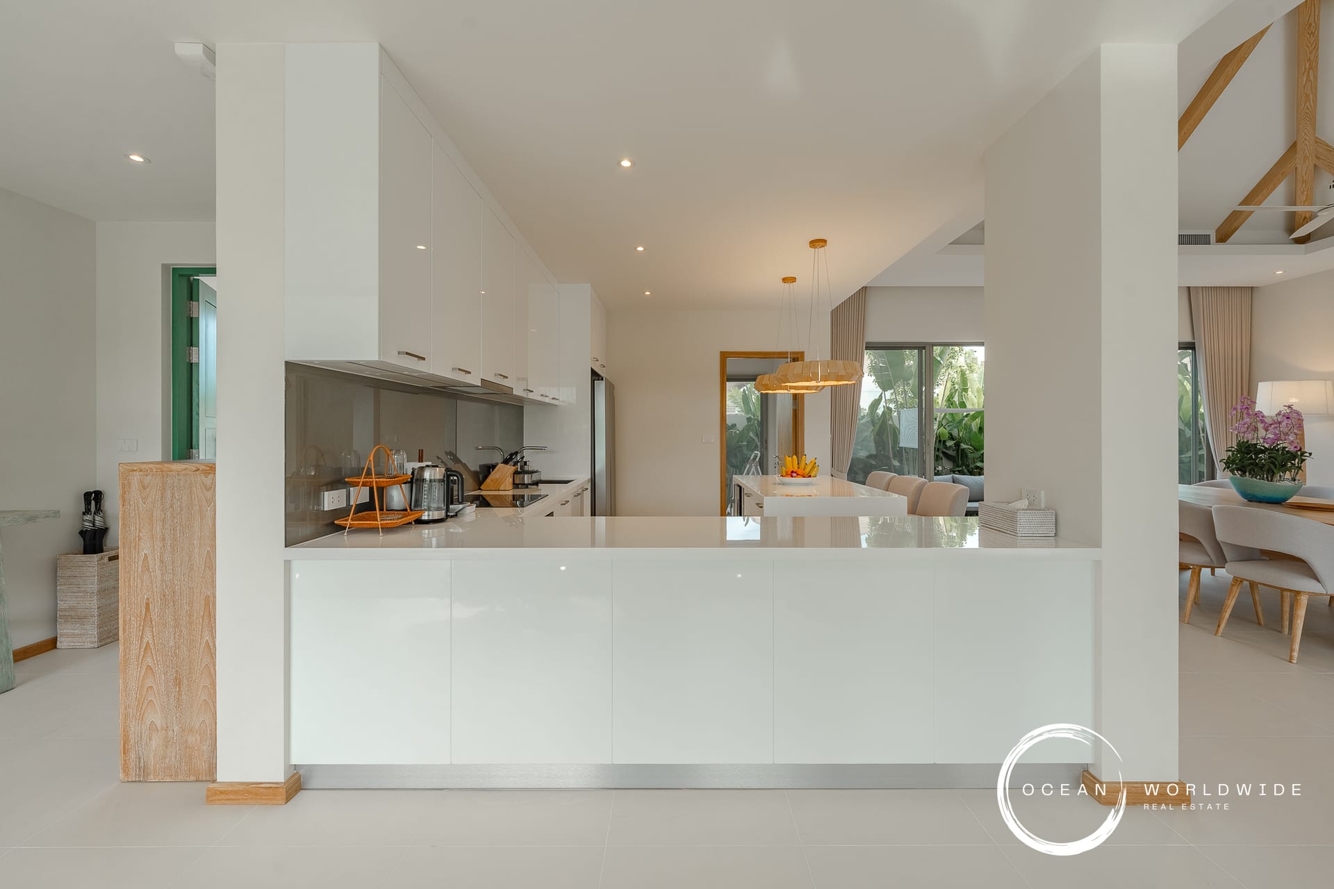 Bright, modern kitchen with white cabinetry and an island, open to a dining area with beige chairs and large windows showing greenery outside.
