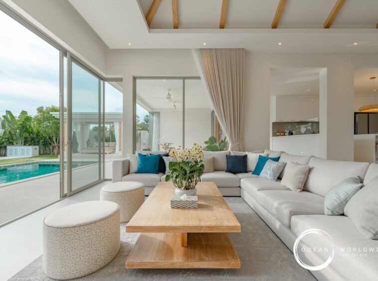 Bright open-plan living room with a large beige sectional sofa, wooden coffee table, and sliding glass doors to a pool area outside.