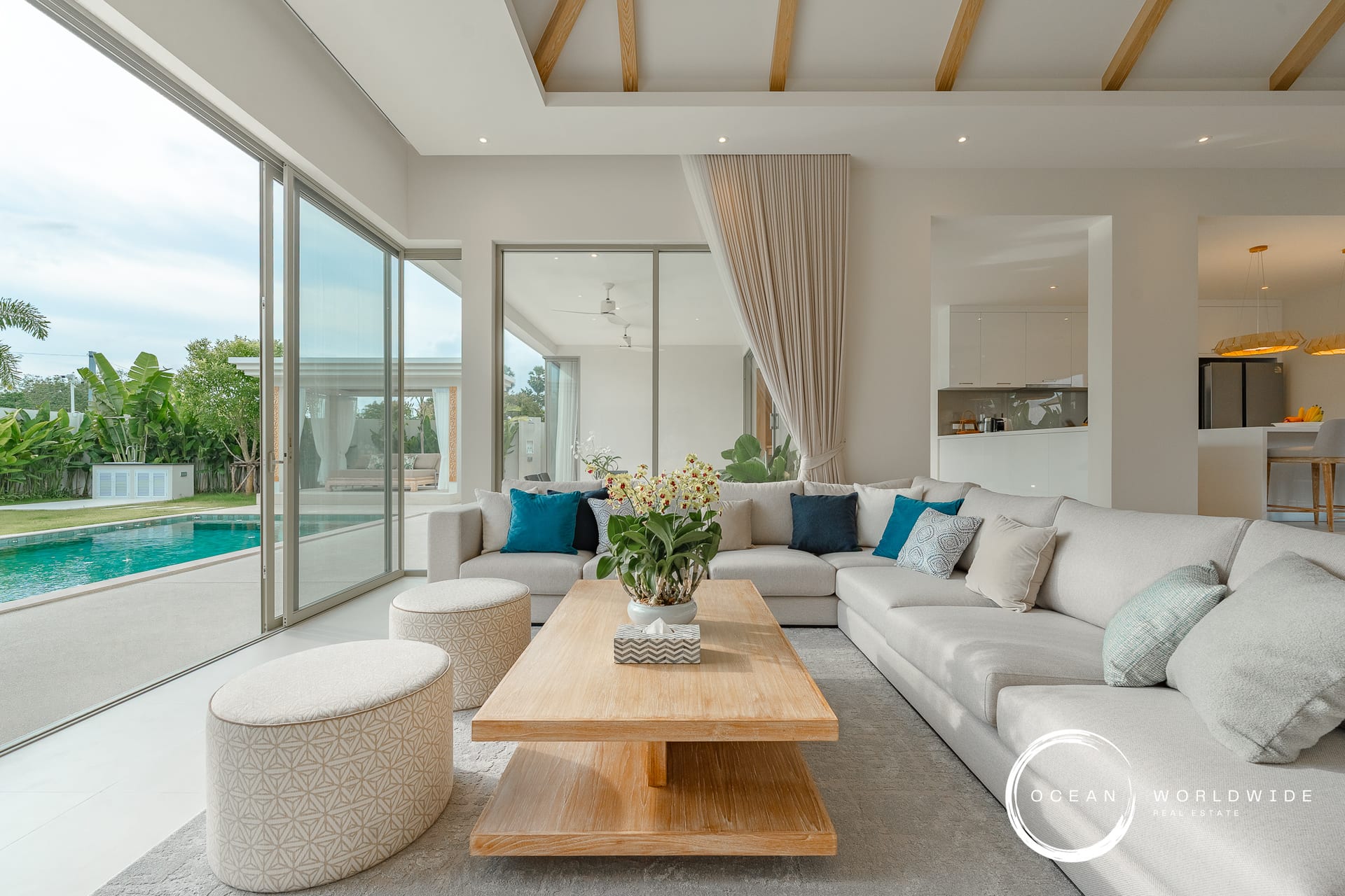 Bright open-plan living room with a large beige sectional sofa, wooden coffee table, and sliding glass doors to a pool area outside.