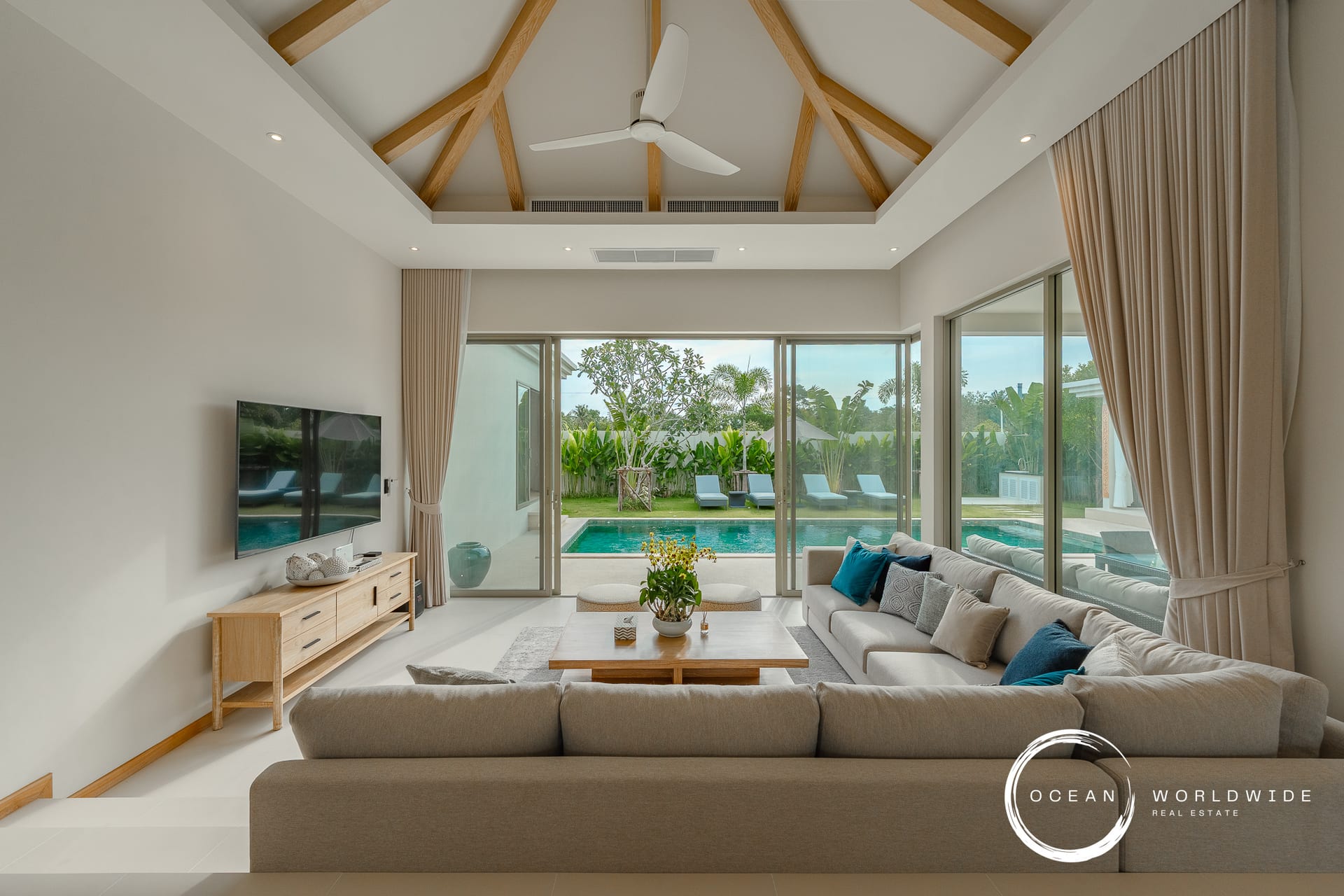 Bright living room with beige sectional facing sliding glass doors to a pool yard, vaulted ceiling with wooden beams and a white ceiling fan.