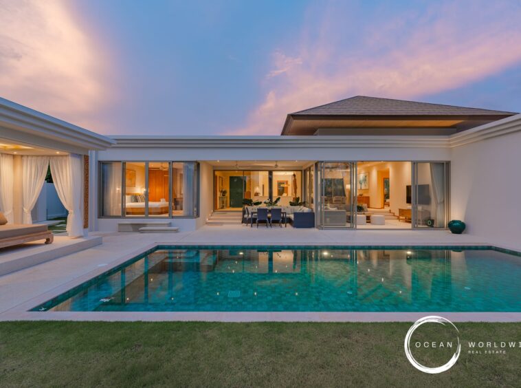 Modern luxury villa with a rectangular turquoise pool, glass walls, and open living spaces at sunset. Ocean Worldwide branding visible.