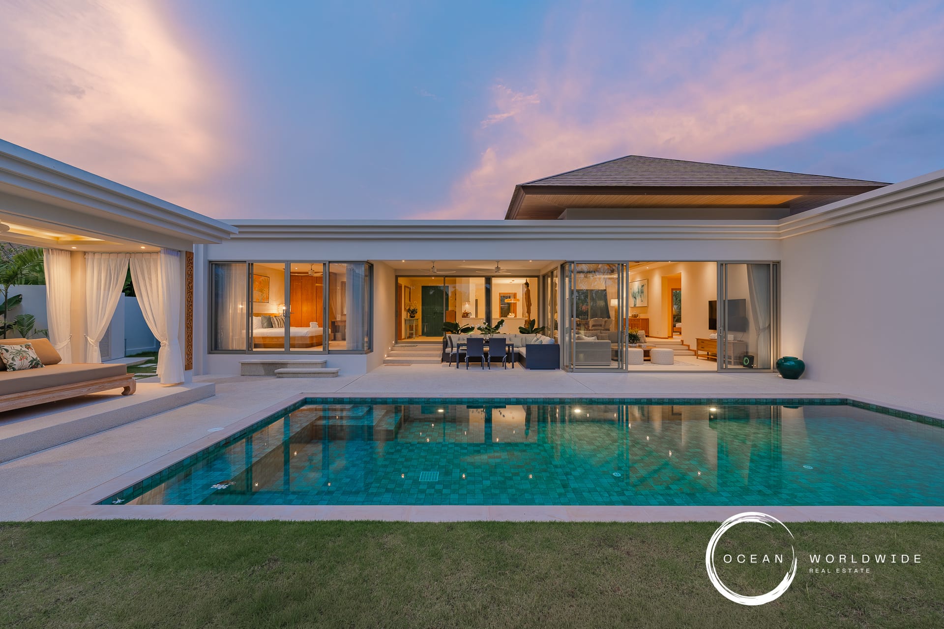 Modern luxury villa with a rectangular turquoise pool, glass walls, and open living spaces at sunset. Ocean Worldwide branding visible.