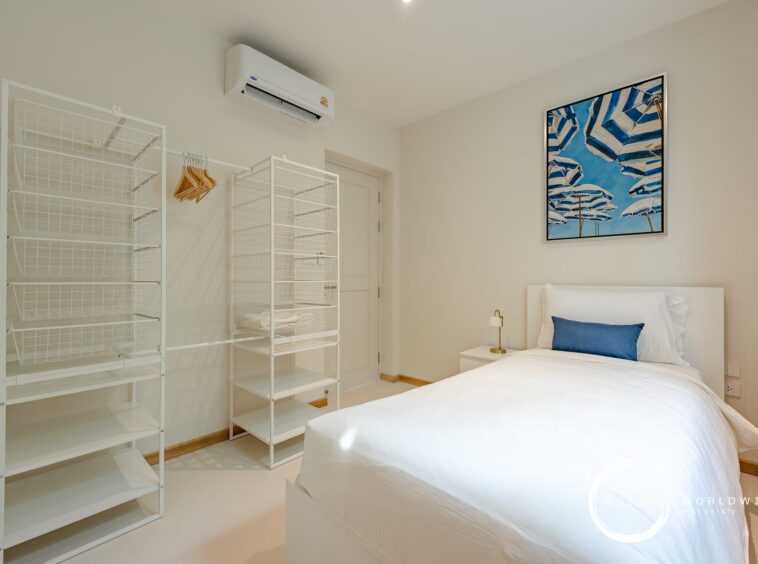 Minimalist white bedroom with two white wire shelving units, a bed with white linens and a blue accent pillow, and a framed blue umbrella photo on the wall
