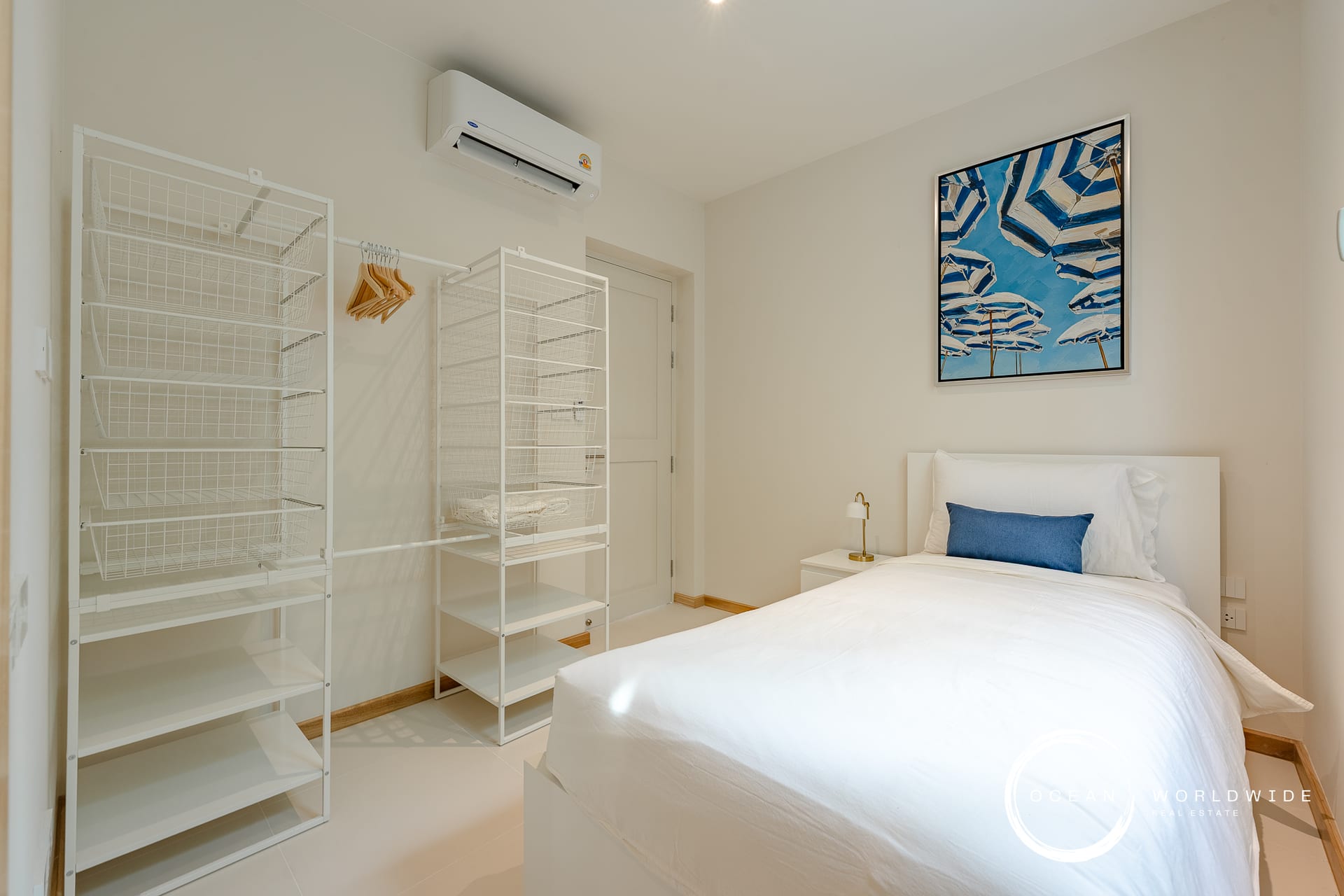 Minimalist white bedroom with two white wire shelving units, a bed with white linens and a blue accent pillow, and a framed blue umbrella photo on the wall