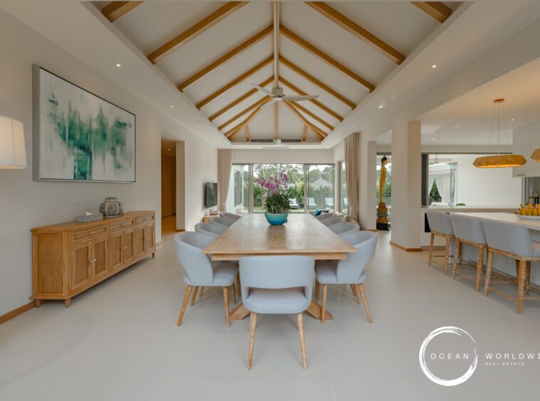 Open-plan dining area with a vaulted ceiling and exposed wooden beams, a long wooden table surrounded by gray chairs, and a kitchen island with pendant lights on the right.