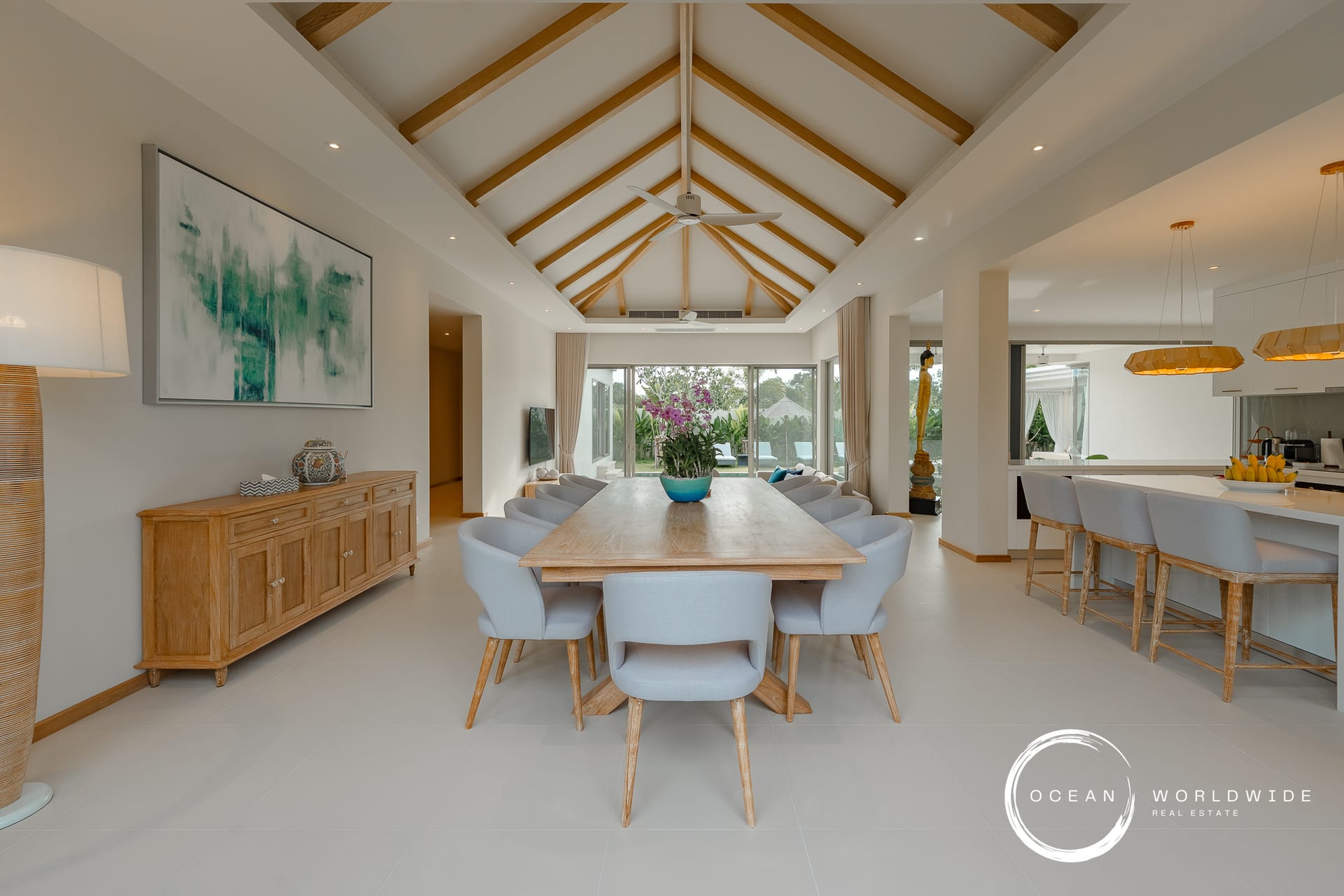 Open-plan dining area with a vaulted ceiling and exposed wooden beams, a long wooden table surrounded by gray chairs, and a kitchen island with pendant lights on the right.