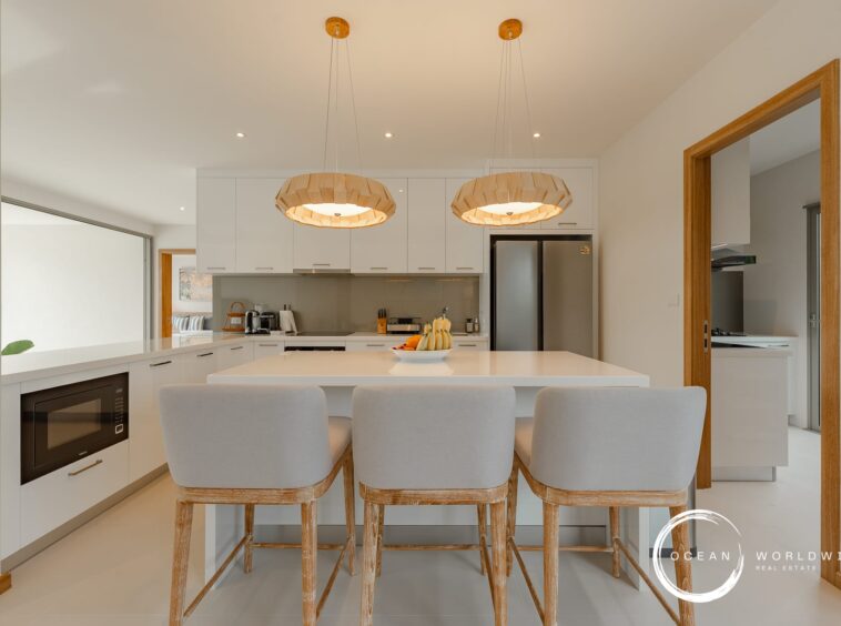 Bright modern kitchen with white cabinets, stainless steel fridge, and a large white island with four wooden stools beneath two pendant lights.