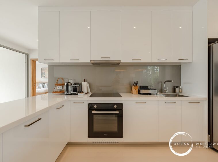 Modern white kitchen with glossy cabinetry, built-in oven, stovetop, and stainless fridge along a white countertop island