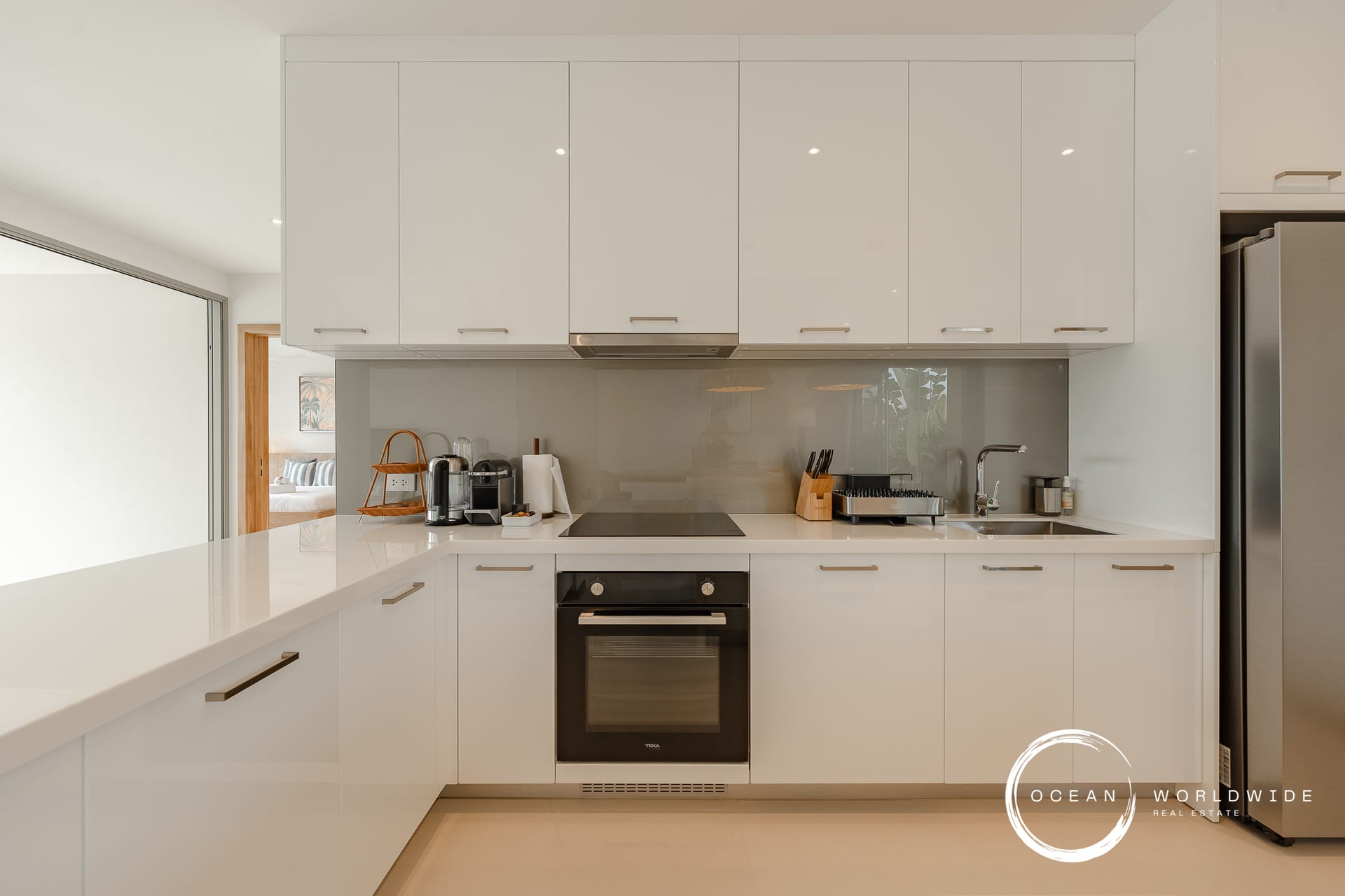 Modern white kitchen with glossy cabinetry, built-in oven, stovetop, and stainless fridge along a white countertop island