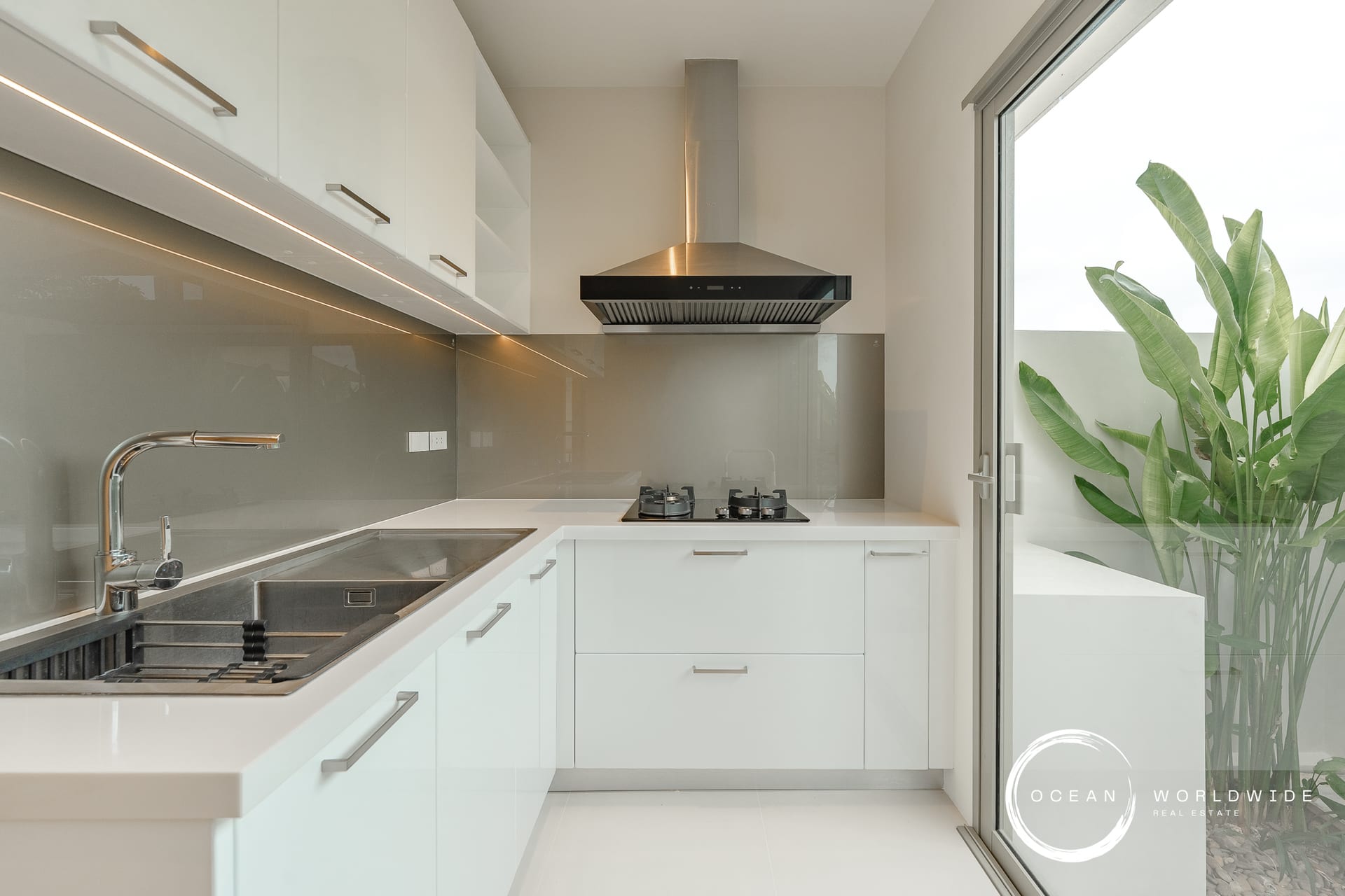 Modern white kitchen with stainless steel range hood, gas cooktop, sink, and a glass door opening to a plant-filled patio on the right.