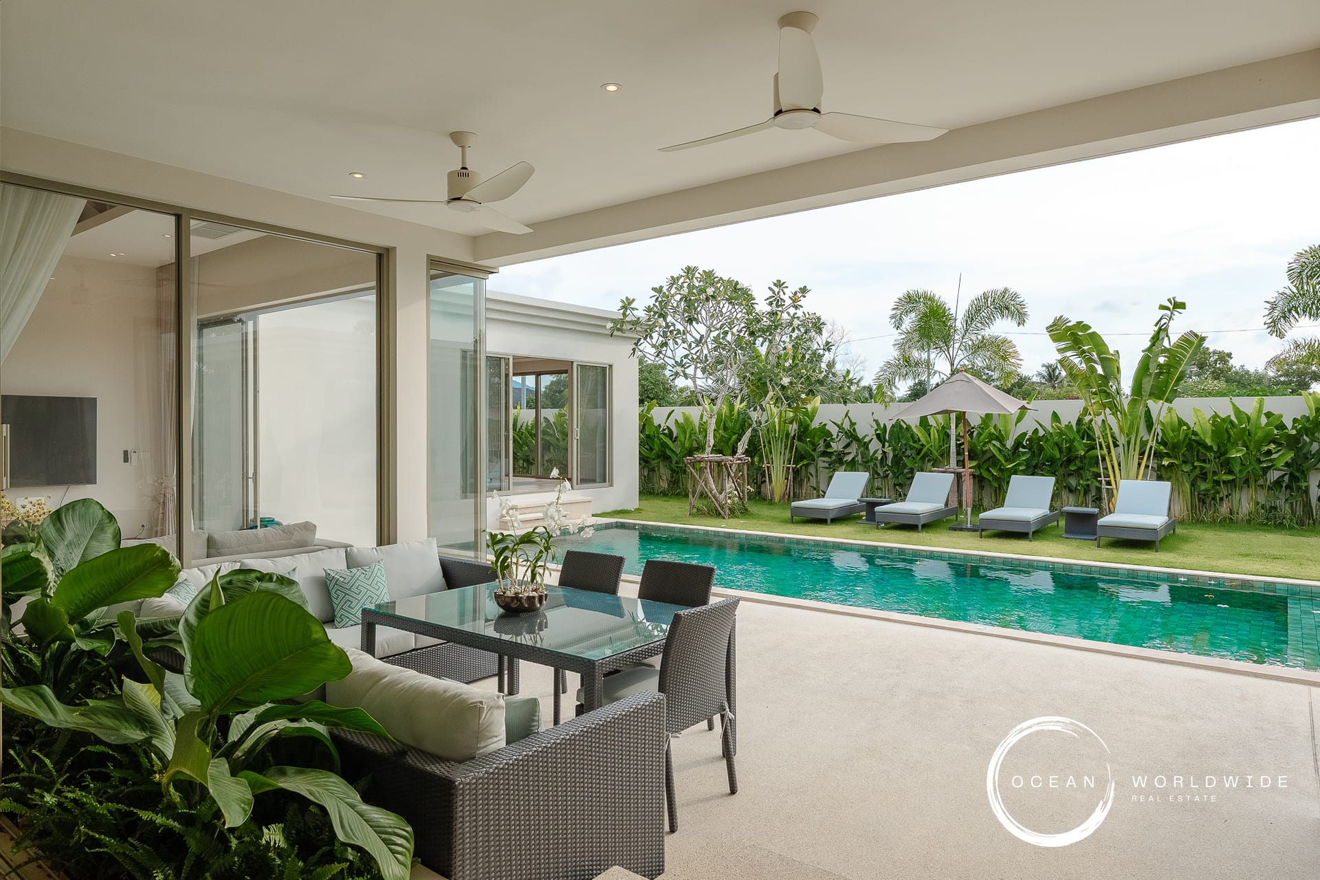 Poolside covered patio with gray wicker seating, tropical plants, and a rectangular turquoise pool.