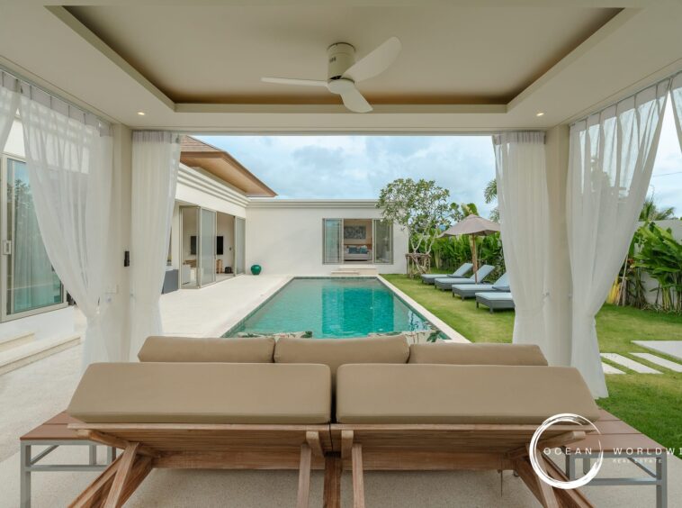 Outdoor living area with beige cushioned seating under a covered patio overlooking a rectangular pool and lawn.