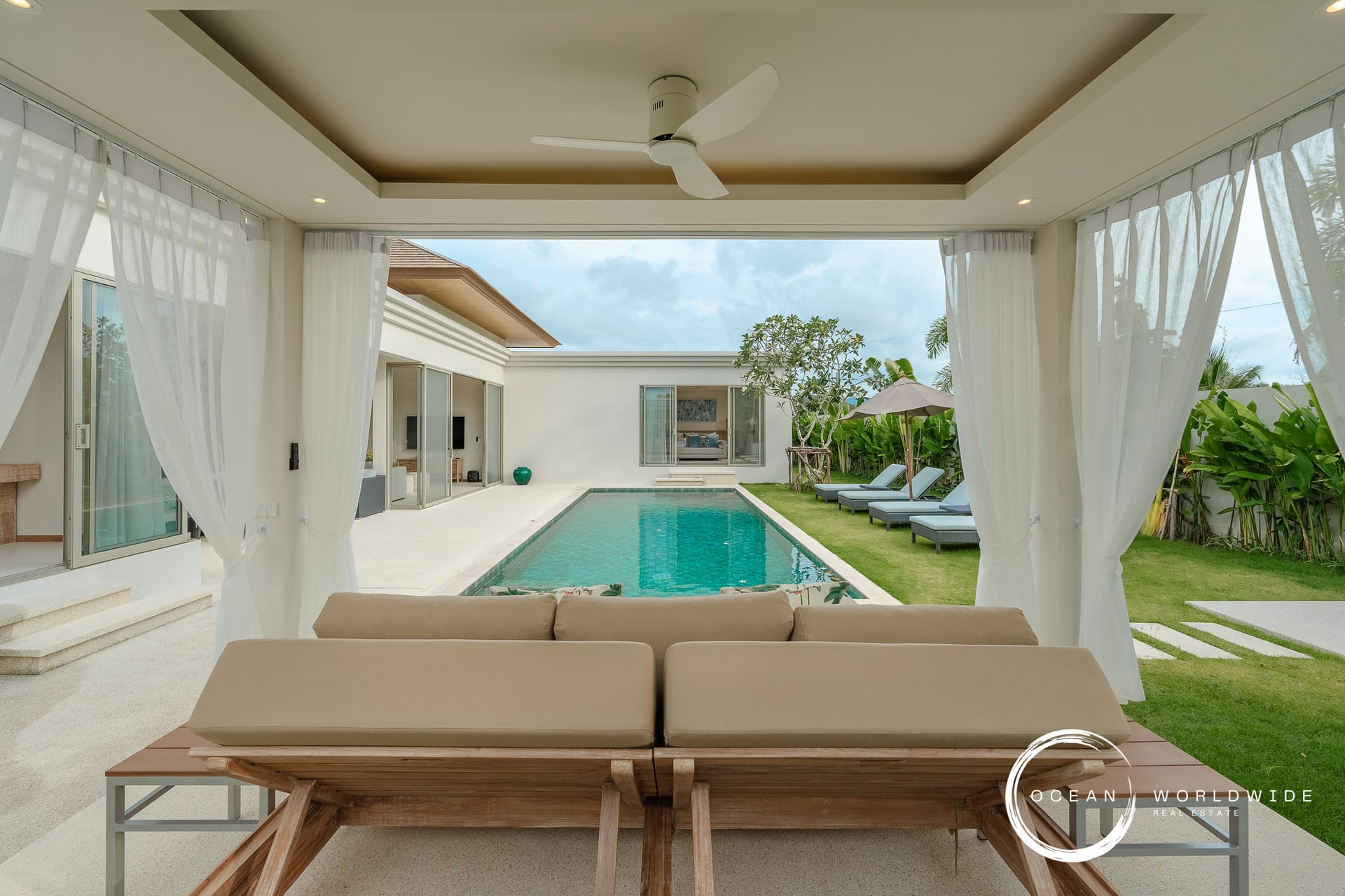 Outdoor living area with beige cushioned seating under a covered patio overlooking a rectangular pool and lawn.