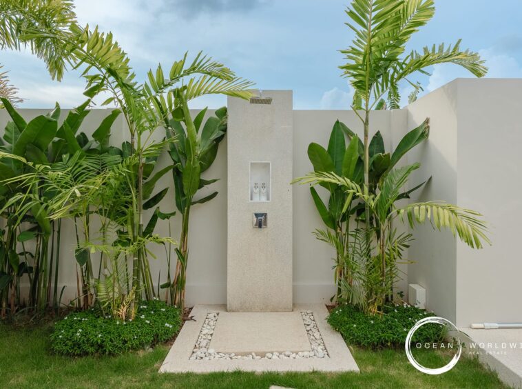 Outdoor shower pillar in a tropical garden, surrounded by tall palm plants and a pebble-bordered concrete path. Watermark bottom right reads Ocean Worldwide Real Estate.