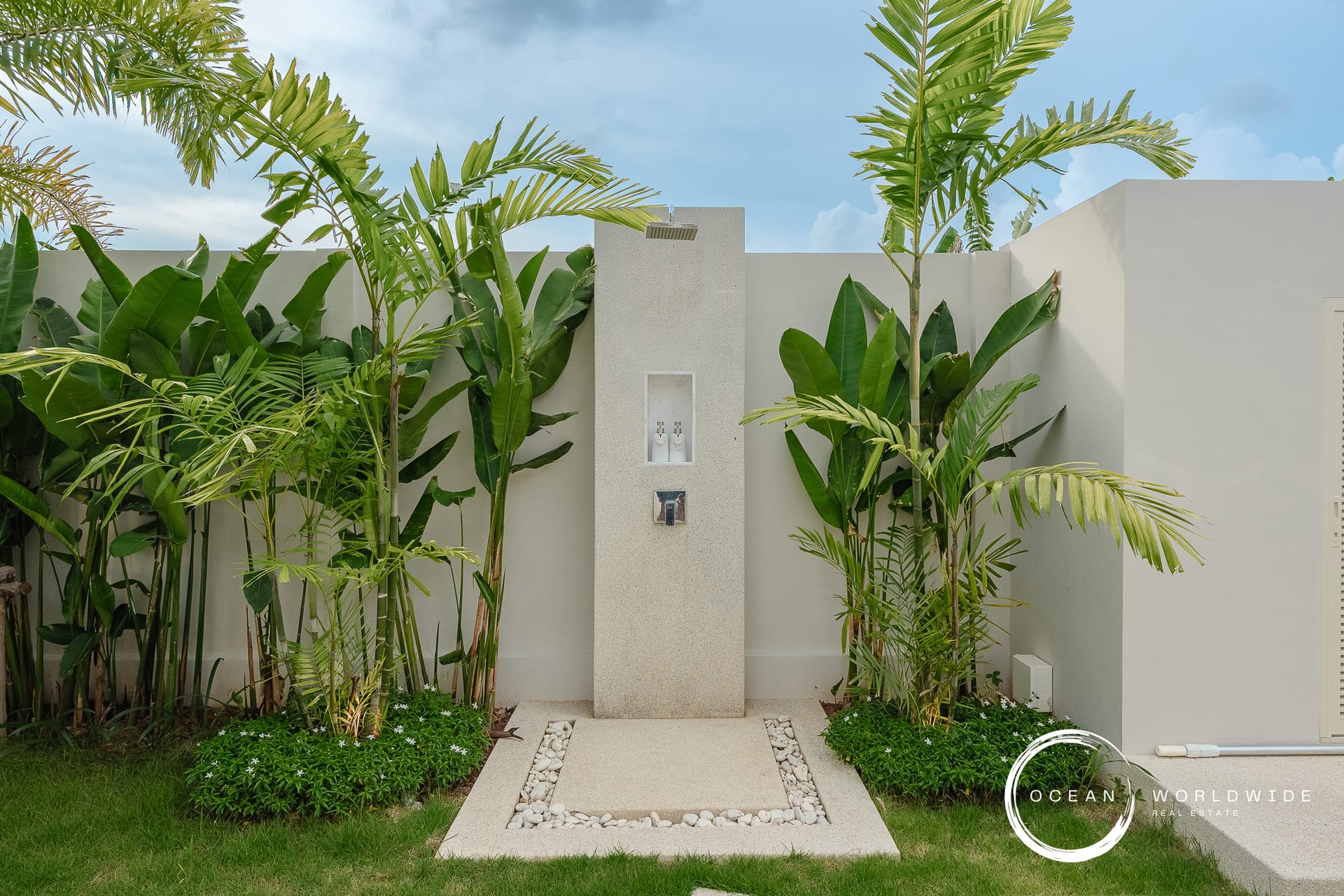 Outdoor shower pillar in a tropical garden, surrounded by tall palm plants and a pebble-bordered concrete path. Watermark bottom right reads Ocean Worldwide Real Estate.