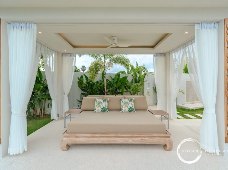 Open-air cabana with white drapes, a beige daybed, and tropical plants creating a resort-like lounging area.
