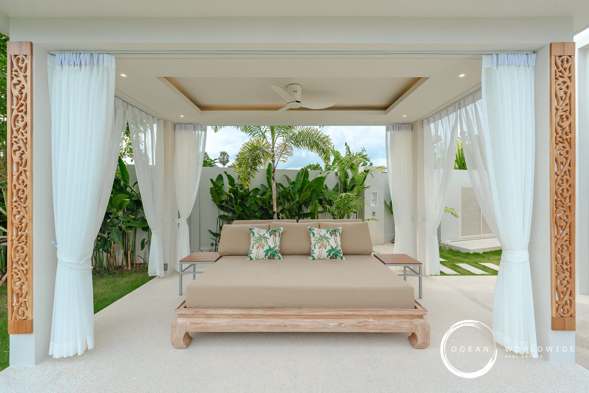 Open-air cabana with white drapes, a beige daybed, and tropical plants creating a resort-like lounging area.