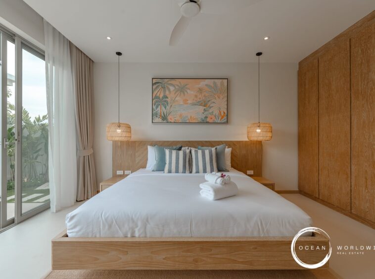Bedroom with a low wooden bed frame, tropical painting, and two woven pendant lamps over a white-tiled floor. Large sliding glass door to a garden.