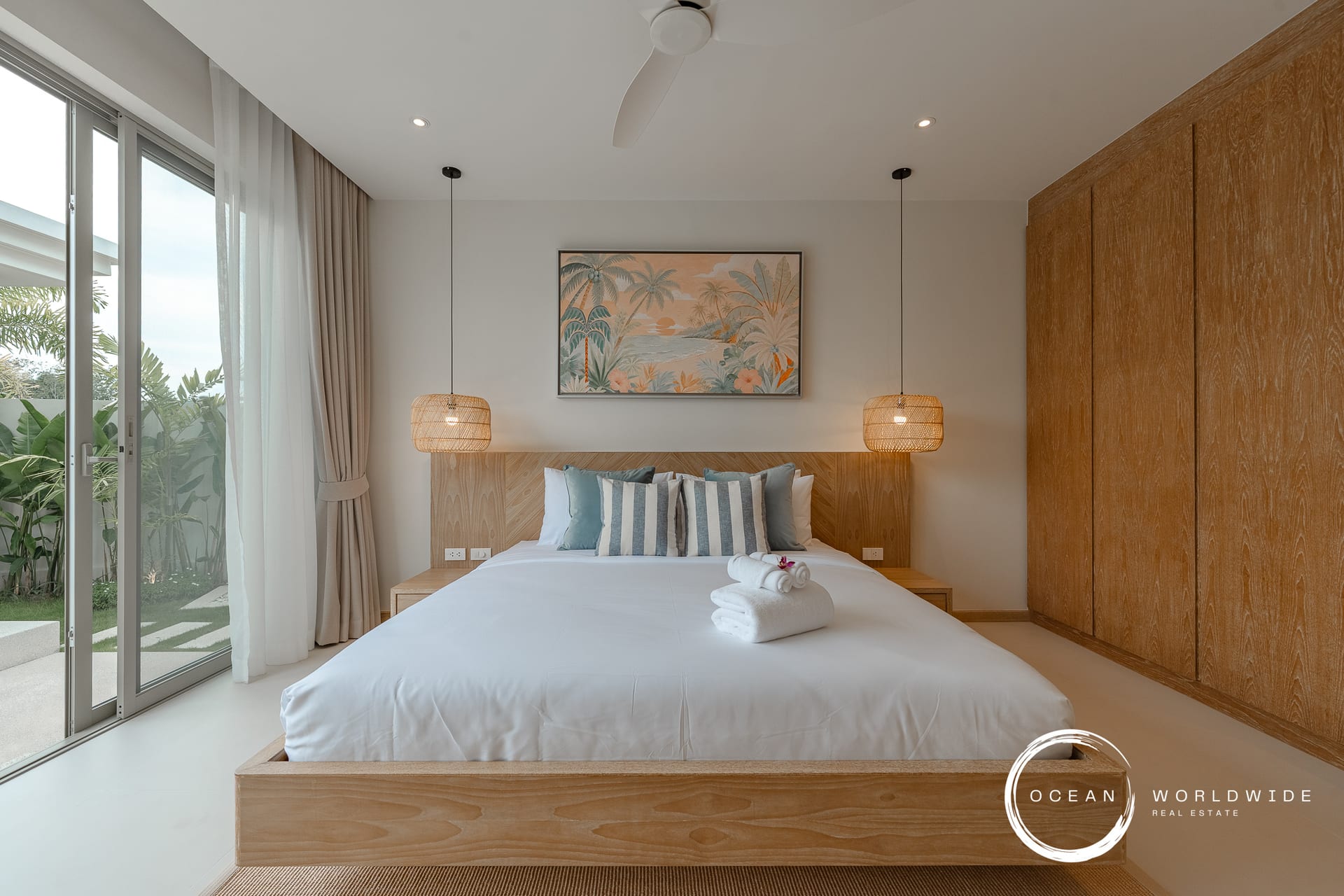 Bedroom with a low wooden bed frame, tropical painting, and two woven pendant lamps over a white-tiled floor. Large sliding glass door to a garden.