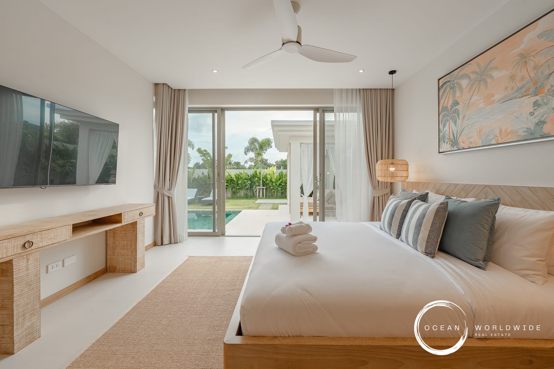 Bright hotel bedroom with a sliding glass door opening to a pool and tropical garden, bed with striped pillows, and a wall-mounted TV on the left.