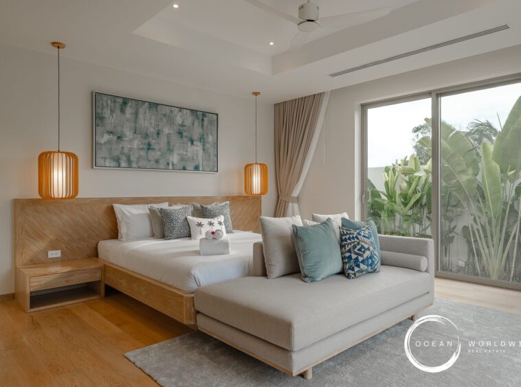 Hotel bedroom with a wooden platform bed, orange pendant lights, abstract wall art, and a gray sofa near sliding glass doors to greenery outside.