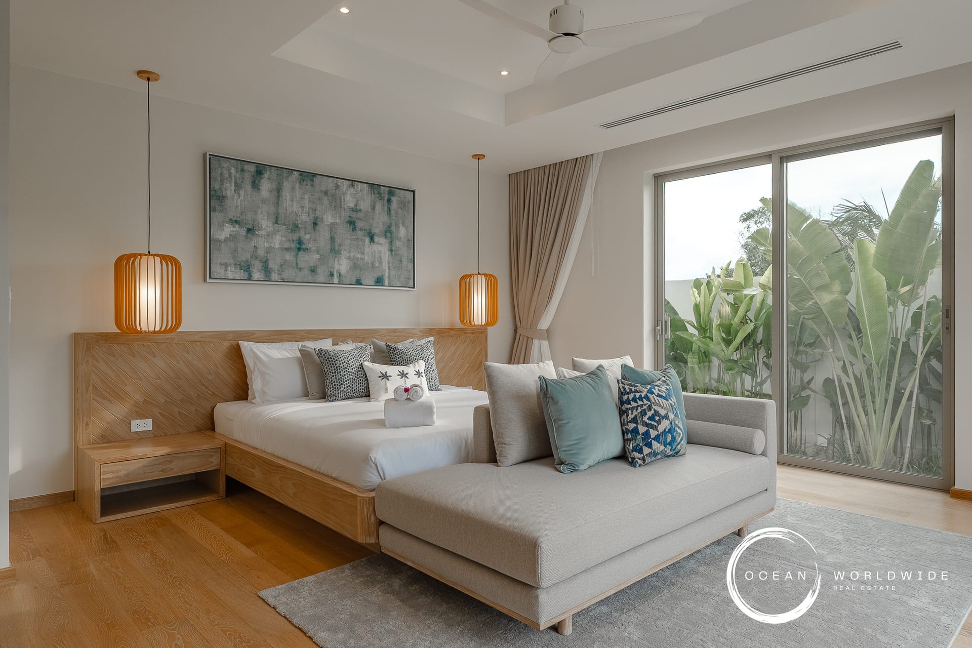 Hotel bedroom with a wooden platform bed, orange pendant lights, abstract wall art, and a gray sofa near sliding glass doors to greenery outside.