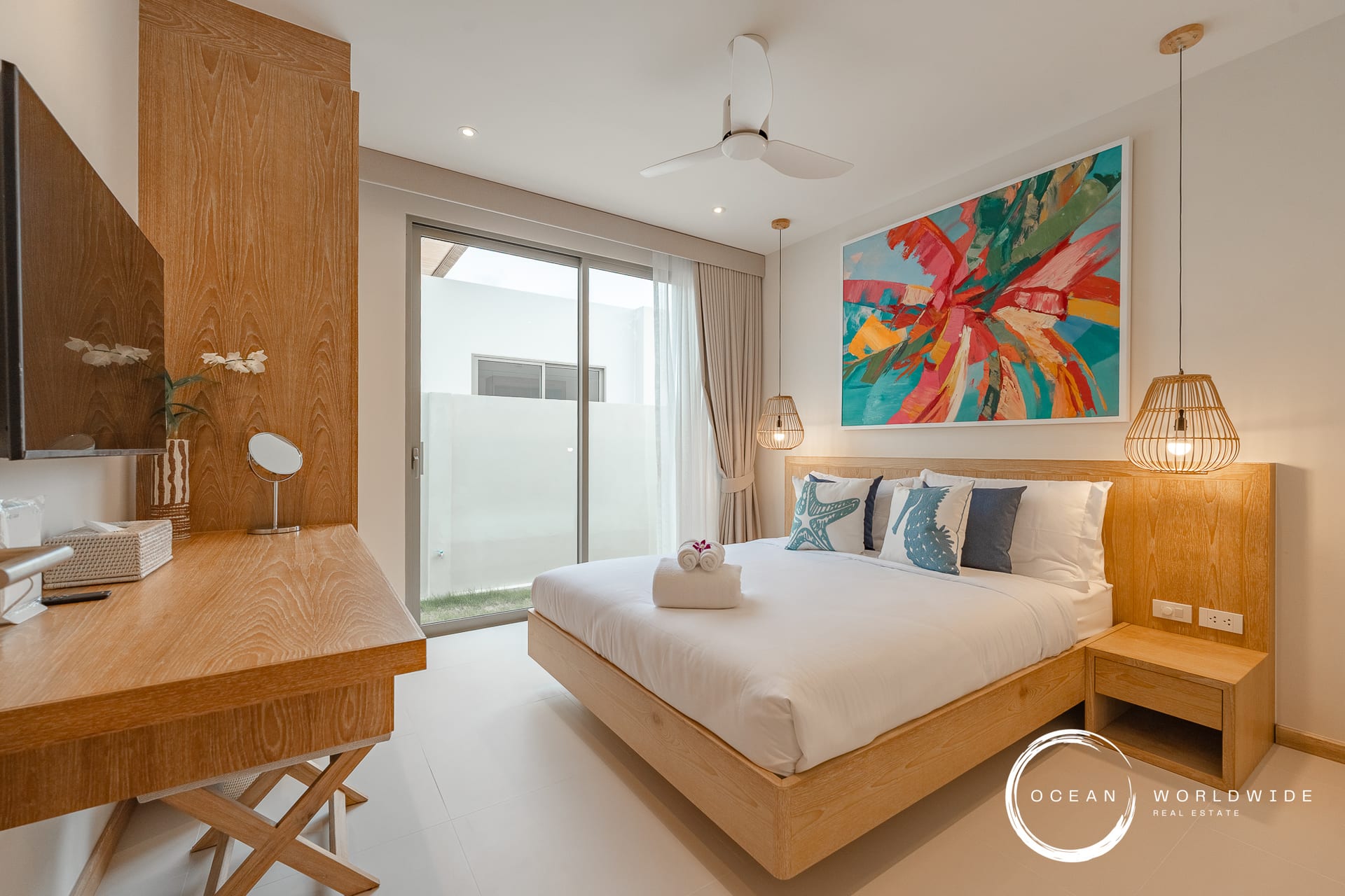 Bright hotel bedroom with a wooden bed frame, abstract colorful wall art, and hanging pendant lights by the nightstands.