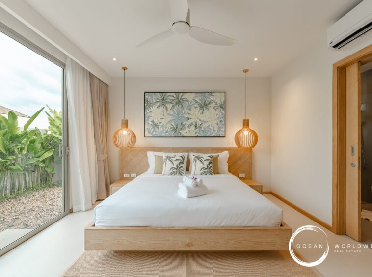 Bright hotel-style bedroom with a low wooden platform bed, tropical palm artwork, and two woven pendant lamps; sliding glass door opens to a lush garden.