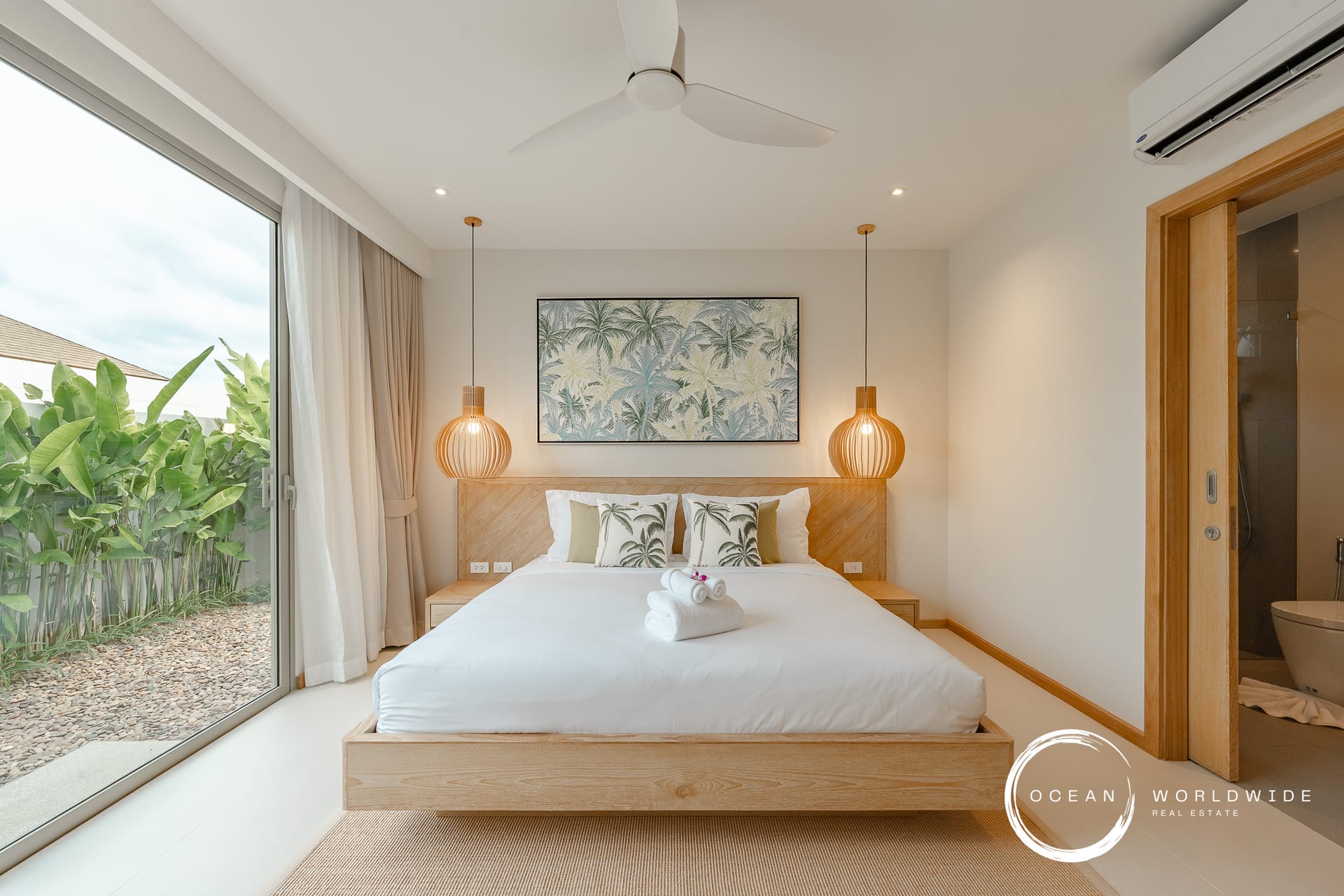 Bright hotel-style bedroom with a low wooden platform bed, tropical palm artwork, and two woven pendant lamps; sliding glass door opens to a lush garden.
