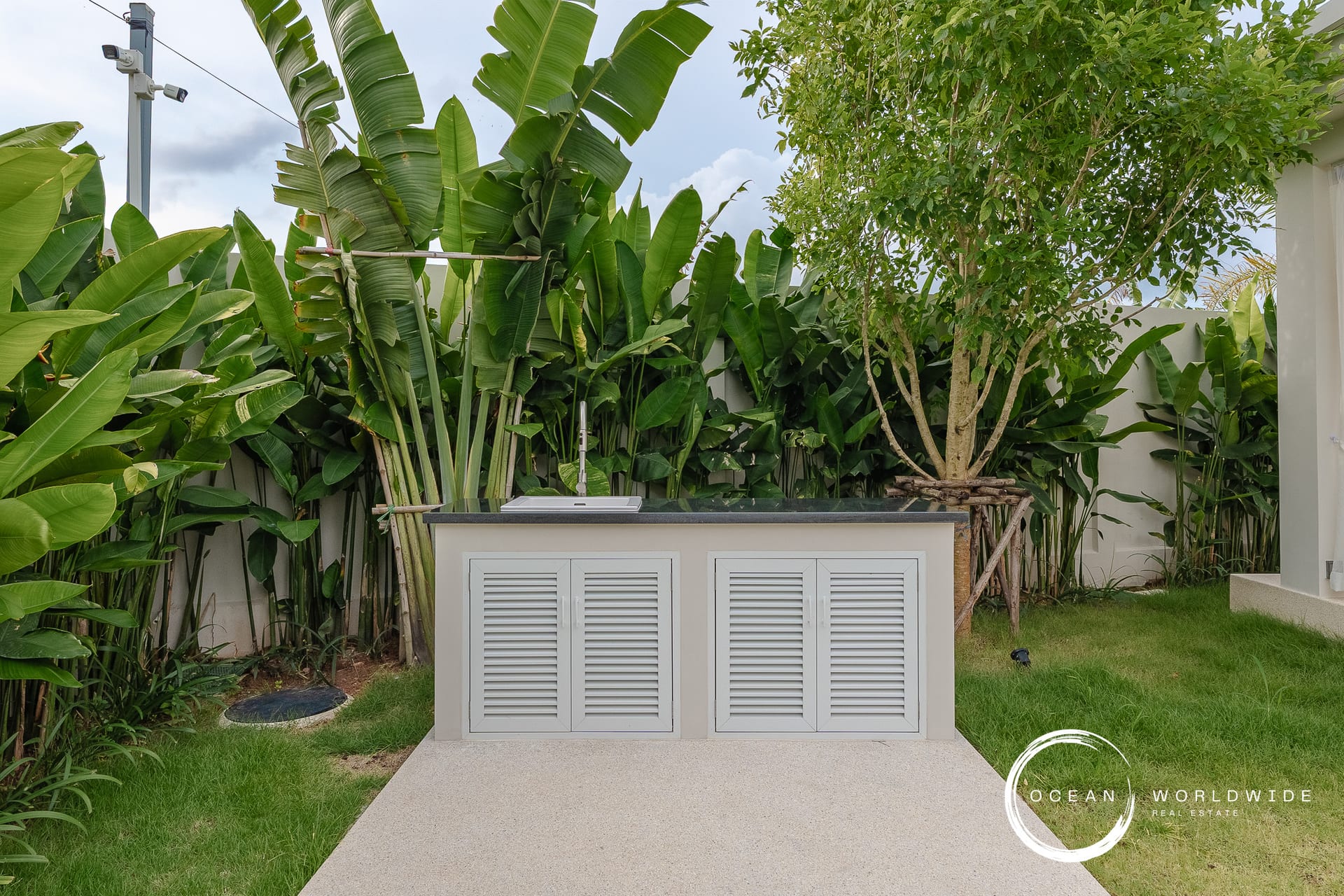 Outdoor utility cabinet with louvered doors set in a tropical backyard behind lush banana plants and a tree.